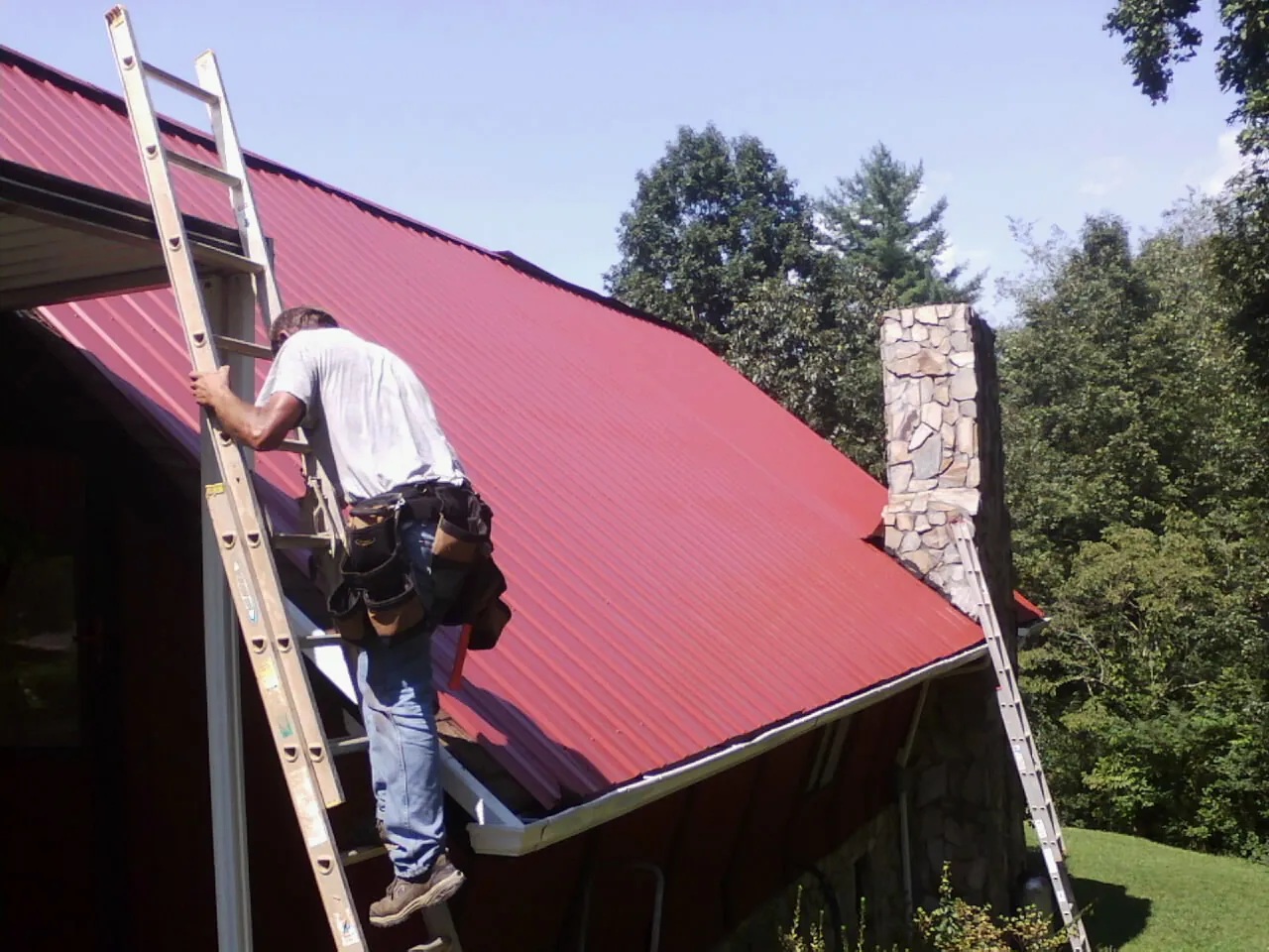 Roofer on a ladder beside a red metal roof and stone chimney — Soffit & Fascia Repair by Roofing Brothers in Ansonia